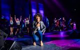 Whitney Houston impersonator in black leather jacket and jeans crouches, smiling at camera as she sings into hand mic, smiling, backing singers and band behind