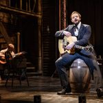 Wild Bill Hickok (Vinny Coyle) sits on a barrel, acoustic guitar resting on his leg as he gazes to the side, singing in an empty saloon, a lone guitar player shadowed in the background