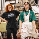 Carrie Hope Fletcher (Calamity Jane) looks at Seren Sandham-Davies (Katie Brown) with her hands on hips as Seren twirls her apron, smiling as she stares upwards