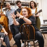 Carrie Hope Fletcher (Calamity Jane) sits backwards on a chair, waving her hands dramatically as the cast crowd around her to listen