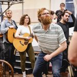 Cast and musicians watch as Man with beard crouches, staring at a figure half out of frame