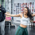 Deena Kapadia in green sweatpants and white strap top dances, arms thrown out