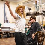 Isaac Savage (Francis Fryer) singing with arm thrown wide wearing blonde curly long haired wig and straw hat, clasping the shirt of Ben Mabberley (Hank & Stage Door Keeper) who sits behind him looking scared and perturbed