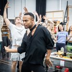 Johannes Radebe dances wearing black, cast dance with arms thrown in air behind him