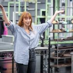 Kara Lily Hayworth, arms in the air as though in bemused question, wearing half tucked in blue and white striped shirt and jeans