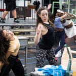 Kaya Farrugia & Kathryn Barnes bend sideways as they dance, bench of shoemaking equipment out of focus before them