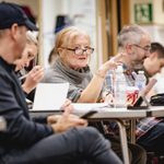 Catherine Jayes (Orchestrations and Music Supervision) gestures from a table with Nick Winston (Co-Director) and other creatives busy with scripts and ipads