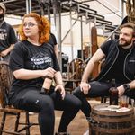 Theo Diedrick (Swing) stands behind Carrie Hope Fletcher (Calamity Jane) and Vinny Coyle (Wild Hill Hickok) who are sitting at a barrel having beer