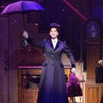 Mary Poppins (Stefanie Jones) holds her umbrella aloft, confetti falling from inside it, inside the hallway, as Mr Banks and the maid watch on.