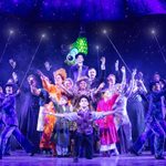 Company of Mary Poppins at night raising hands to the sky. In the centre, Mary Poppins the children, Bert, the bird lady, on the outskirts shimmering chimney sweeps holding up poles with lights on the end. In the sky behind them the parrot of Mary Poppins umbrella hangs