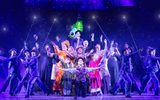 Company of Mary Poppins at night raising hands to the sky. In the centre, Mary Poppins the children, Bert, the bird lady, on the outskirts shimmering chimney sweeps holding up poles with lights on the end. In the sky behind them the parrot of Mary Poppins umbrella hangs