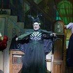 Mrs Brill (Rosemary Ashe) screaming over a handful of red flowers, Miss Andrew (Wendy Ferguson) dressed in black and white blouse, black cape and skirt and large black headpiece, and Winifred Banks (Lucie-Mae Sumner) looking dubiously at her from the staircase.