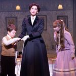 Mary Poppins (Stefanie Jones) with bored expression, holds out a tape measure for Michael (Charlie Donald) to inspect as Jane (Florence Swann) watches