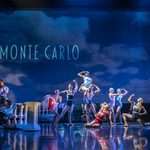 Sign for Monte Carlo in cloudy sky above old-style swimsuit wearing crowd posing, with two beachballs. To the left, by a stone railing two men in shirts relax.