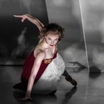 Victoria Page (Cordelia Braithwaite) doing a push up on one hand, staring the camera down, wearing red bodice and white skirt