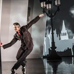 Grischa Ljubov (Glenn Graham) in red and black pinstripe suit mid dance with legs crossed, presenting a red ballet shoe. An old triple lamppost and silhouette of a town behind him