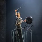 Julian Craster (Stephen Murray) stands on a stage light platform, beckoning with one hand, conductors baton raised in to other