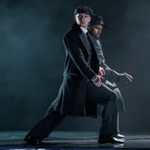 Thomas Shelby in black coat and peaked cap lunges, hand on his leg with a finger cocked, a black man mirroring him behind