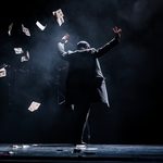 Figure in black coat and peak cap faces away in the darkness, hands thrown up, paper money floating down from them