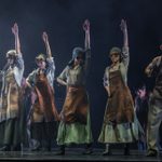 Six workers in leather aprons and round goggles stand in a line, one hand on hip, the other raised in the air