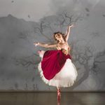 Victoria Page (Ashley Shaw) in white tulle skirt, with red velvet over dress standing en pointe on one foot, other leg raised behind her and arms up in foggy, windy forest, trees bare of leaves