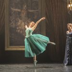 Victoria Page (Ashley Shaw) wearing green dress and shoes, dancing en pointe, one leg raised behind her, arms thrown up, in room with large oil portrait and evening wear people watching