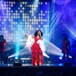 Black woman in red and white ruffled dress sings on purple lit stage with white uplights and two red sequin dress wearing backing singers