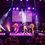 Tribute The Jackson 5 band in gold flared trousers, yellow shirts and red waistcoats sing in front of disco ball screen