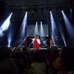 Black woman in red and white ruffle dress sings on stage lit by white spotlights with band behind her