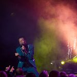 Black man in dark teal suit passionately clenches fist as he kneels, singing, before an audience on a smoky stage