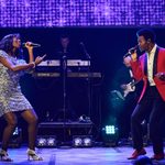 Black woman in silver sequin dress and black man in red jack sing into hand mics before screen with purple sequins on