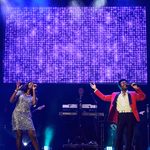 Black woman in silver sequin dress and black man in red jack sing into hand mics before screen with purple sequins on