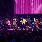 Black man in white jacket sings at black woman in silver sequin dress as others dance around and band plays behind them