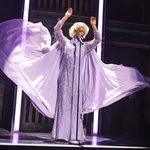 Lola (Johannes Radebe) in long lilac dress with flowing cape and blonde wig, singing into standing mic, arms artistically in the air