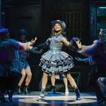 Lola (Johannes Radebe) in denim cowgirl dress and hat in the spotlight, dancers in shadow surrounding