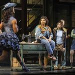 Lauren (Courtney Bowman) in denim sits on a desk with a sewing machine staring along with factory workers as Lola (Johannes Radebe) in denim cowgirl dress and hat prances in