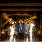 Blonde woman in black and gold dress stands on a podium, other black and gold wearing dancers stand around her as four bursts of fire shoot up front of stage