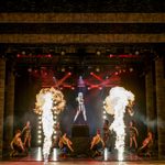 Blonde woman in black and gold dress stands on a podium, other black and gold wearing dancers look up at her as four bursts of fire shoot up front of stage