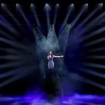 Brunette woman sings on stage, lights pointing in from every angle