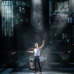 Charlie (Dan Partridge) in white vest, reaches upward in spotlight in the shoe factory