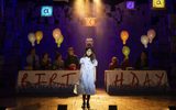 Young girl in blue and white dress and glasses holds pile of books in string holder as birthday party happens behind her with balloons, a cake raised aloft and a birthday banner across the tables