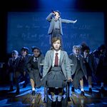 School girl sits on chair staring ahead, boy stands singing on table behind her as others dance around