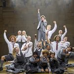 School children jubilantly crowd around, posing towards the camera as a small boy singing into microphone, fist in the air, is raised amongst them