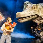 Female ranger holds up an egg with a baby Giraffatitan emerging to a large puppet Giraffatitan