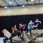 Line of male dancers on tilted stage reach outwards