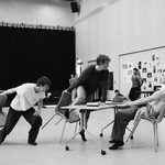 041125_SadlersWells_Quadrophenia_REHEARSAL-1-0094 Black and white image of two men titling chairs forward toward a book laden desk where a third man is sitting, leaning away