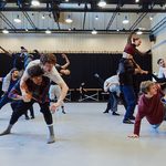 To the left, a bent double man gives another a piggy-back, to the right, a man stretches out on the floor, behind dancers support each other in tall multi-people lifts
