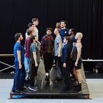 Two rows of dancers stand on a tilted stage, a man in plaid walking downslope between them