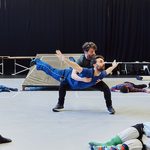 A man holds another man at knee height, the man in blue has his arms out like he is soaring through the air, bodies scatter the ground around them