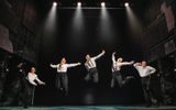 Five people in white shirts jumping gleefully on dark stage lit by four spotlights